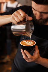 Young smiling male barista making cappuccino. Pouring milk for prepare cup of coffee. Latte art. Morning cup of coffee in café. Brewing coffee. Coffee shop concept. - obrazy, fototapety, plakaty