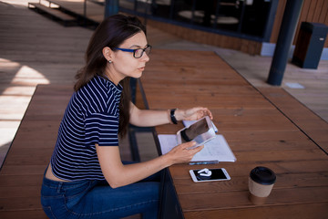 A serious brunette girl, freelancer, with large wrist watch, works at a wooden table on tablet,...