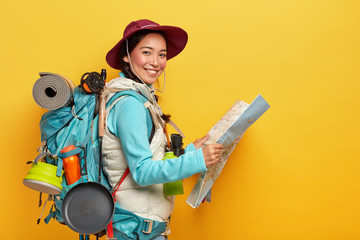 Active korean female tourist carries big rucksack, wears hat and casual clothes, holds map, studies...