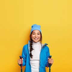 Vertical image of happy active Asian female stands with trekking poles, wears blue hat and jacket, being on mountain trail, hikes during autumn vacation in forest, isolated over yellow background