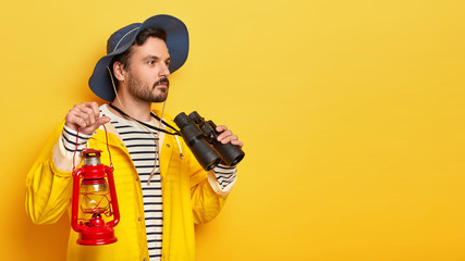 Profile shot of serious unshaven man vacationist holds gas lamp, observes something into distance with binoculars, wears hat and raincoat, poses over yellow background. People and camping concept