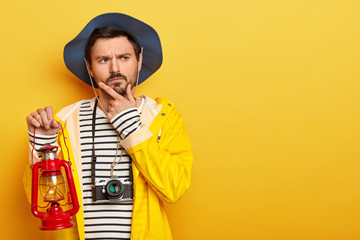 Photo of thoughtful man holds chin, thinks about having journey or expedition, holds little gas lamp, dressed in raincoat, headgear, uses camera for making photos, isolated over yellow background