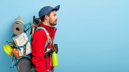 Sideways shot of thoughtful unshaven man wears hat, red jacket, carries backpack with necessary things, covers long distance on foot, has adveture trip, poses against blue background, copy space