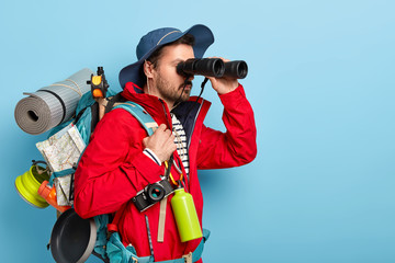 Trekking, camping, hiking concept. Photo of serious male tourist uses binoculars to observe...