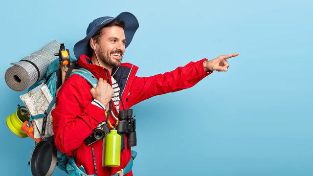 Pleased younng male traveler has hiking trip, wears hat and casual jacket, carries backpack on shoulders, points into distance with index finger, stands against blue background. People, trekking