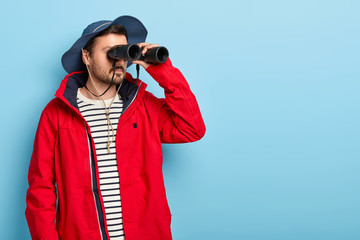 Photo of serious male camper has long adventurous journey, keeps binoculars near eyes, wears hat...