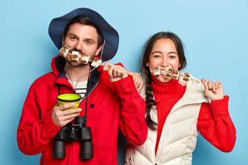 Photo of female and male tourists eat tasty marshmallow roasted on bonfire, spend pastime in nature, like traveling and having adventures, wear casual clothes. Two friends have pinic in forest