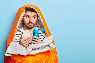 Portrait of male hiker wrapped in sleeping bag, looks thoughtfully away, holds mobile phone and...