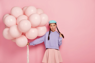Pensive pretty girl with rouge cheeks, dressed in fashionable clothes, holds bunch of balloons, comes on party, thinks what interesting to do to entertain guests, models against pink background