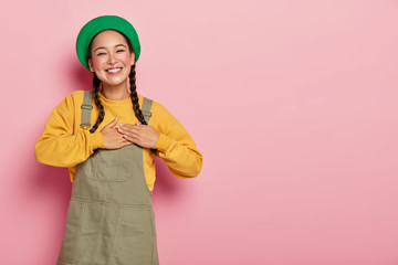 Happy mixed race woman with pigtails has appreciated expression, keeps palms on heart, expresses...