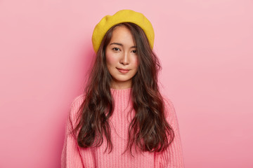 Photo of beautiful brunette woman with mixed race appearance, wears yellow beret and pink knitted sweater, poses against rosy background, looks calmly at camera, enjoys free time for good rest.