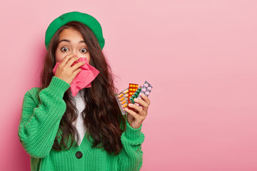 Sick Asian woman caught cold during wet autumn day, has running nose, holds various pills for curing disease, wears green beret and sweater, has ill look, copy space against pink background.