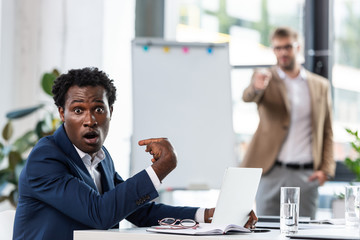 shocked african american businessman sitting at table and pointing with finger at himself in office