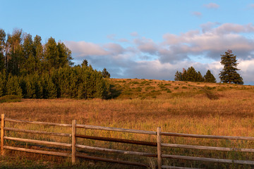 Evening light on wood fence and pasture with trees on hilltop, sky with clouds, Eastern Washington State, USA