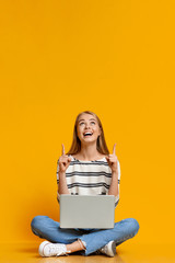 Excited teen girl with laptop pointing upwards at free space