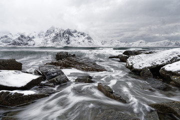 Küstenlandschaft im Winter mit Wasserbewegung zwischen großen Steinen, auf denen Schnee liegt, Wellen laufen auf die Küste zu, im Hintergrund eine Bergkette mit Schnee - Location: Norwegen, Lofoten
