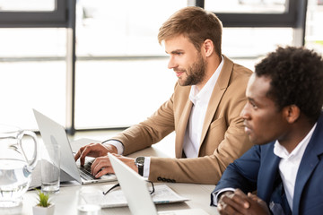 two multiethnic businessmen using laptops at table in office