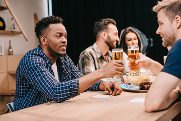 multicultural friends clinking glasses of light beer while spending time together in pub