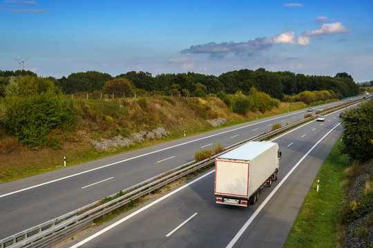 White Truck Is Driving On The Highway Through The Country Landscape Under A Blue Sky With Clouds, Transport And Environment Concept, Copy Space