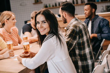 attractive young woman smiling at camera while sitting with multicultural friends together in pub