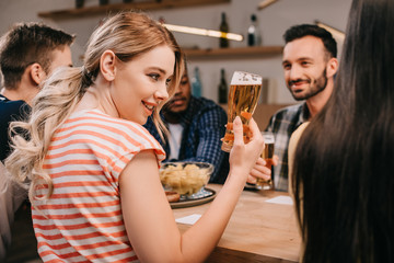 selective focus of smiling young woman holding glass of beer while sitting with multicultural friends in pub