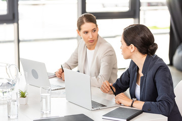 two businesswomen sitting at table with laptops and looking at each other