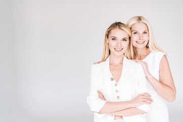 smiling blonde mature mother embracing young daughter in total white clothes isolated on grey