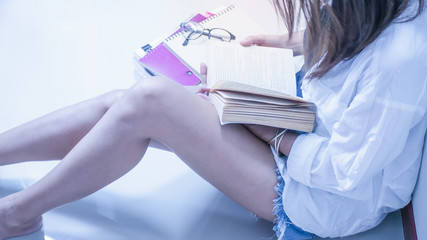 Women wearing white shirts, shorts  , Sitting on a white chair reading a book. And there are stacks of books nearby. Concept  Education,  love to read.