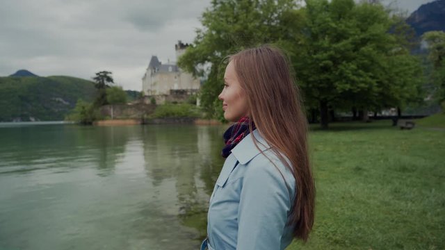 Close up of attractive girl standing in profile on the shores of Lake Annecy. She is wearing blue coat and looking forward with smile. Castle Ruphy is on background. Duingt, France