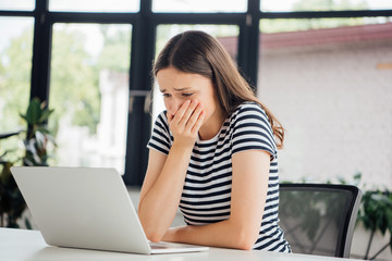 sad girl in striped t-shirt covering mouth with hand while using laptop at home