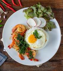 Spicy fried shrimp with basil and chili, serve with rice and egg in white ceramic plate on wood table, Thai style food.