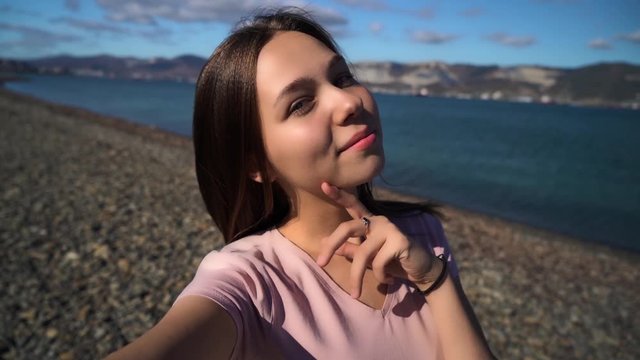 Attractive young woman using mobile and takes selfies on the beach with mountains and cloudy sky on the background. Mobile phone camera view.