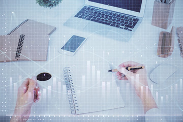 Hands writing information about stock market in notepad. Forex chart holograms in front. Concept of...