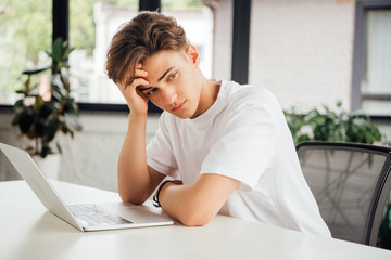 pensive teen boy in white t-shirt sitting at table with laptop and looking away at home
