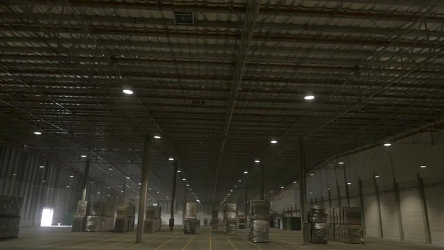 Aerial drone shot: flying inside an anonymous warehouse, among ready-to-ship pallets of goods, under many lamp lights.