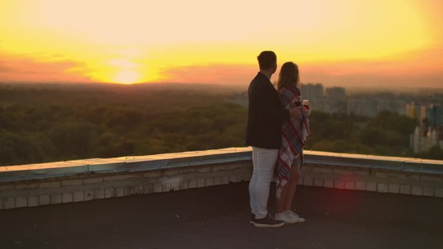 A young couple on the roof drinking wine from glasses standing in a blanket and admiring the beautiful sunset over the city. Romantic evening on the roof overlooking the city.