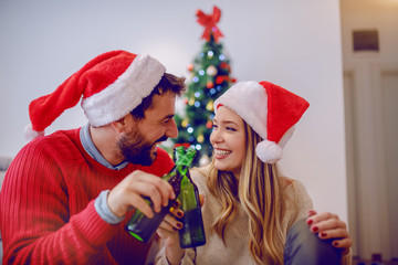 Cheerful handsome caucasian couple with santa hats on heads sitting on the floor and toasting with beer. In background is christmas tree. Living room interior.