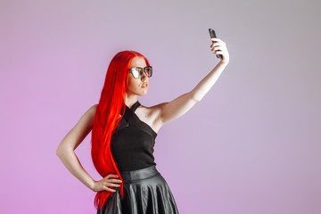 A girl with red long hair wearing glasses and a leather skirt takes a selfie on a pink background