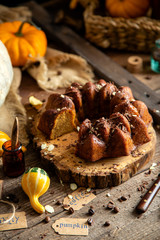 tasty homemade baked pumpkin bundt cake with chocolate on top stands on wooden board on rustic table with assorted colorful small pumpkins and autumn leaves in straw basket