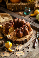 tasty homemade baked pumpkin bundt cake with chocolate on top stands on wooden board on rustic table with assorted colorful small pumpkins and autumn leaves in straw basket