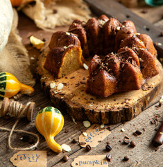 tasty homemade baked pumpkin bundt cake with chocolate on top stands on wooden board on rustic table with assorted colorful small pumpkins and autumn leaves in straw basket