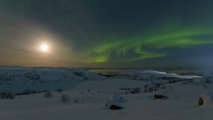 Northern lights, aurora in the sky at night. Hills and rocks covered with snow. Moon in the sky.