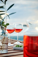 two glasses of rose wine with  olives against blue water on a pier..