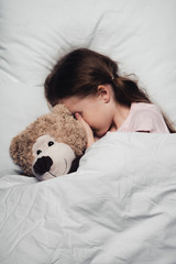 adorable child covering eyes with hand while lying in bed with teddy bear