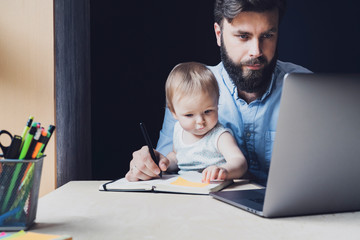 Man and little baby sitting in front of laptop. Serious and busy father with small child on his knees trying to work remotely.