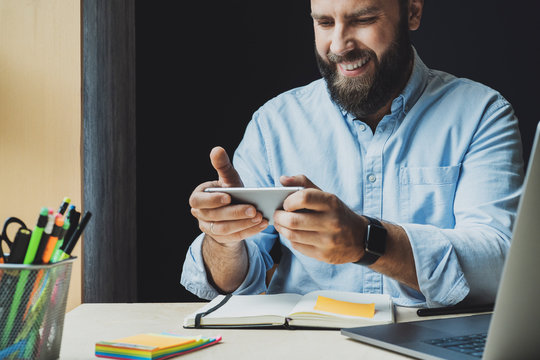 Smiling guy sitting at desk and playing online games on smartphone. Manager spending break time on communicating in social networks. Guy using mobile phone for watching funny videos and communicating