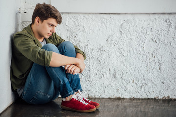 sad teenager in jeans holding smartphone and sitting on floor