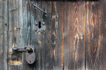 A rusty padlock hangs on a wooden door. The old door from the boards is locked. Rusty handle and rusty lock on a closed wooden door.