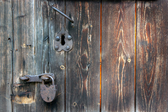 A rusty padlock hangs on a wooden door. The old door from the boards is locked. Rusty handle and rusty lock on a closed wooden door.