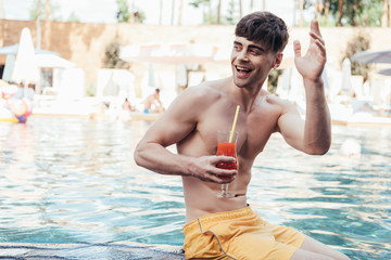 cheerful young man looking away and waving hand while relaxing at poolside with glass of refreshing beverage